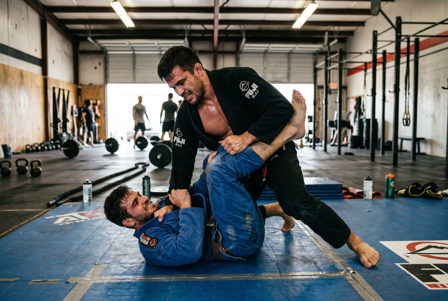 Two BJJ athletes grappling on blue mats in an industrial gym during intense training