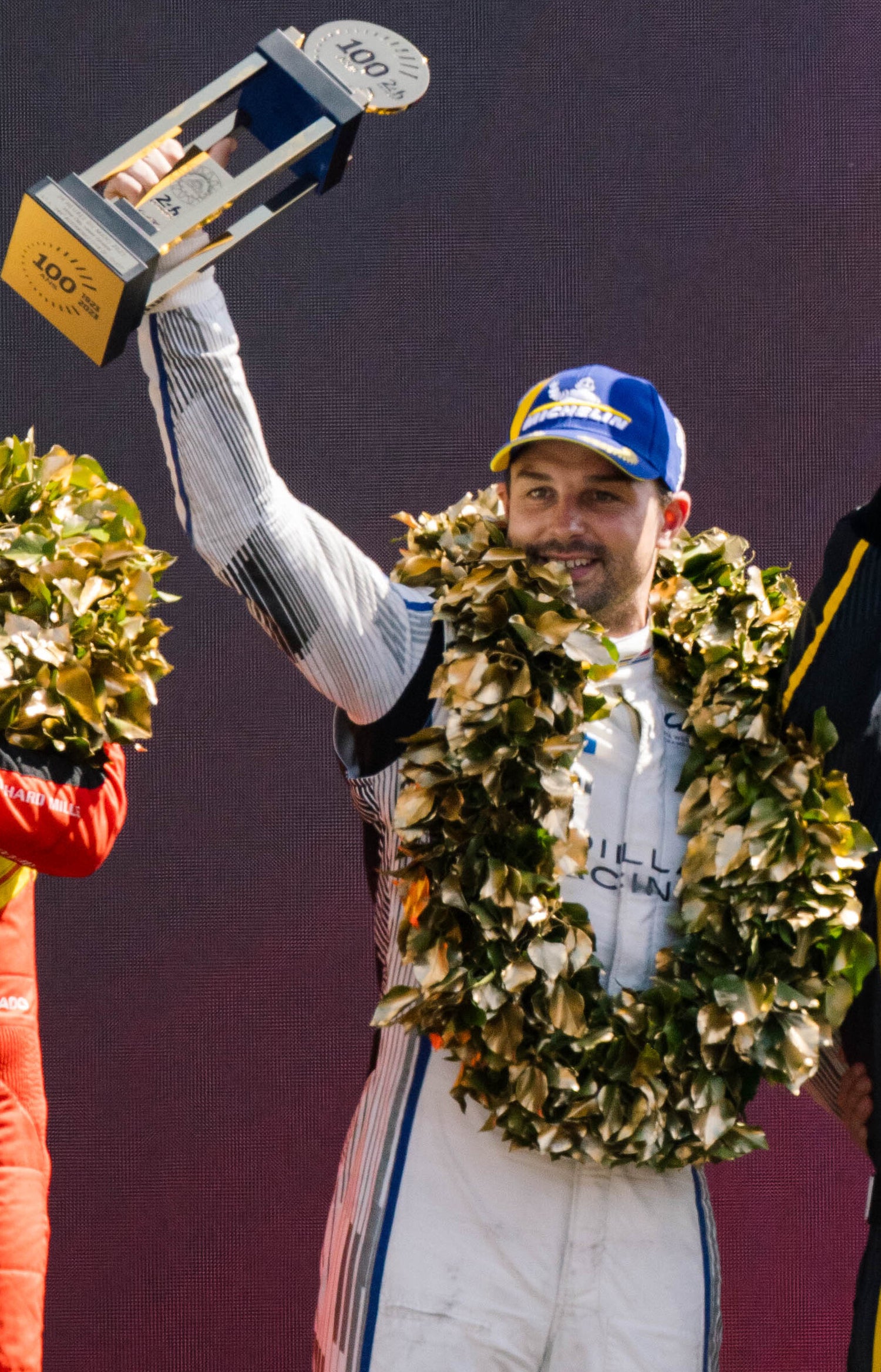 Earl Bamber in racing suit holding a trophy and wreath on a podium